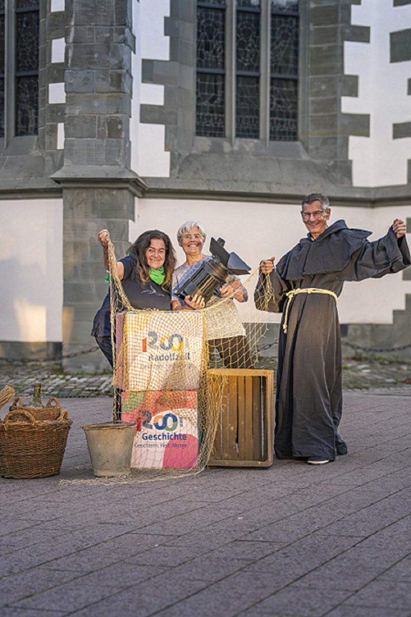 Radolfzell: Gruppe mit Requisite vor Kirche. Mönch, Scheinwerfer, Kisten, Netz. 1200 Jahre Geschichte.