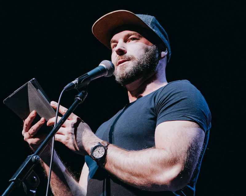 Mann mit Bart und Kappe am Mikrofon. Vortrag, Präsentation, Bühne.