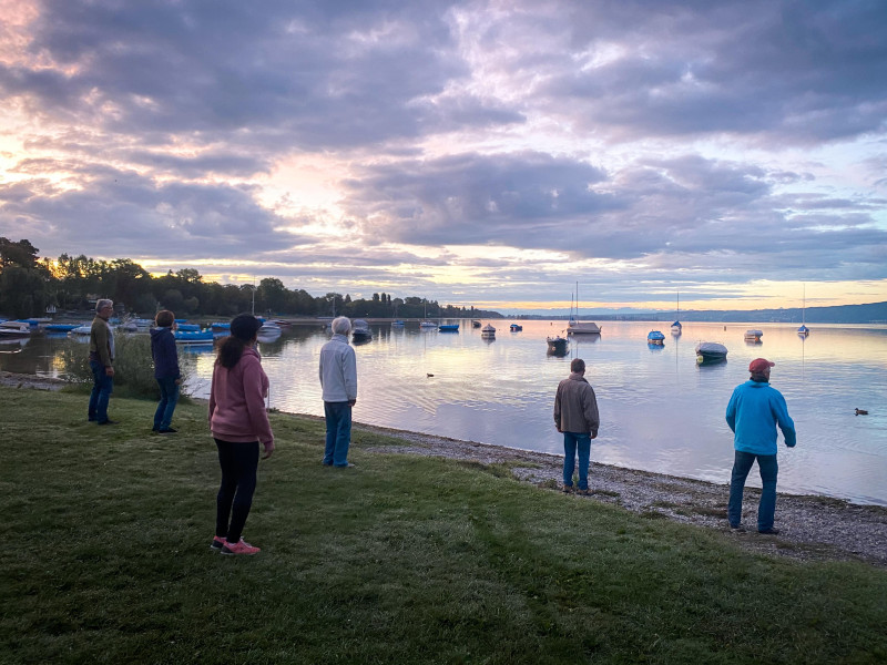 Gruppe von Menschen am Seeufer bei Sonnenuntergang. Boote auf dem Wasser.