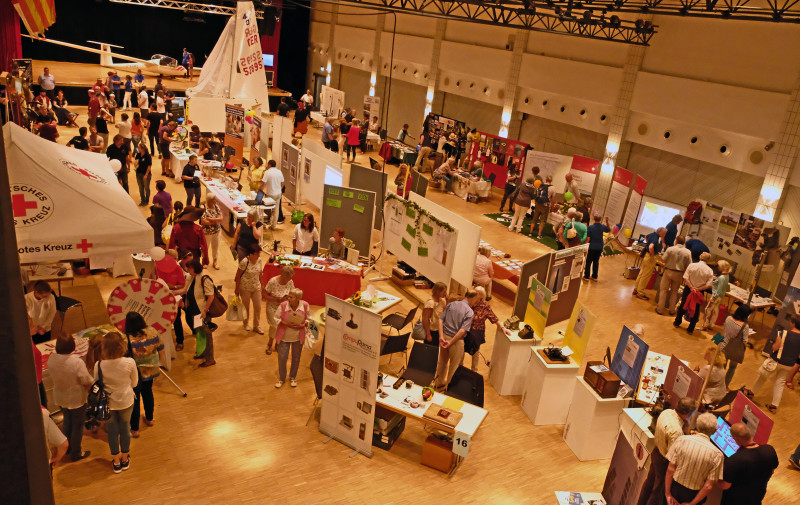 Messe-Überblick: Menschen an Ständen in Halle. Rotes Kreuz Zelt, Segelboot, Flugzeug. Besucher erkunden Ausstellung.